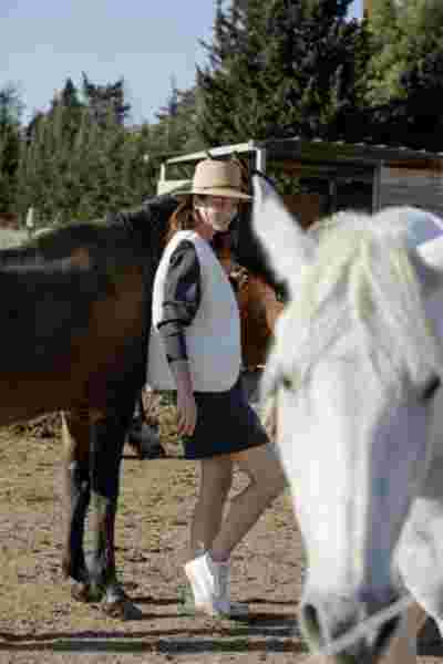 Modèle en robe grise et gilet ecru dans un box, cheval blanc au premier plan — Lookbook April & C, Occitanie