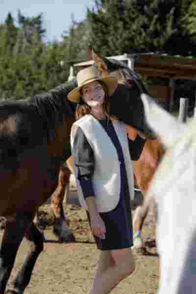 Modèle en robe grise, gilet écru et chapeau de paille appuyée contre un cheval brun — Lookbook April & C, Occitanie